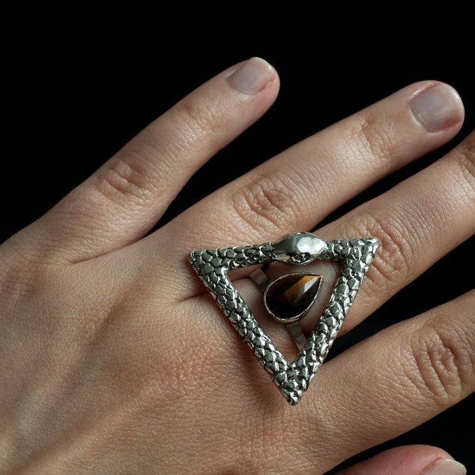Close up of a hand wearing a hand carved snake ring in the shape of a triangle ouroboros, surrounding a pear-shaped tiger eye.