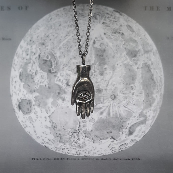 Silver necklace in the shape of a hand with an eye in the palm, also called a hamsa. Shown on a background of the moon. 