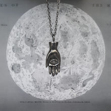 Load image into Gallery viewer, Silver necklace in the shape of a hand with an eye in the palm, also called a hamsa. Shown on a background of the moon. 