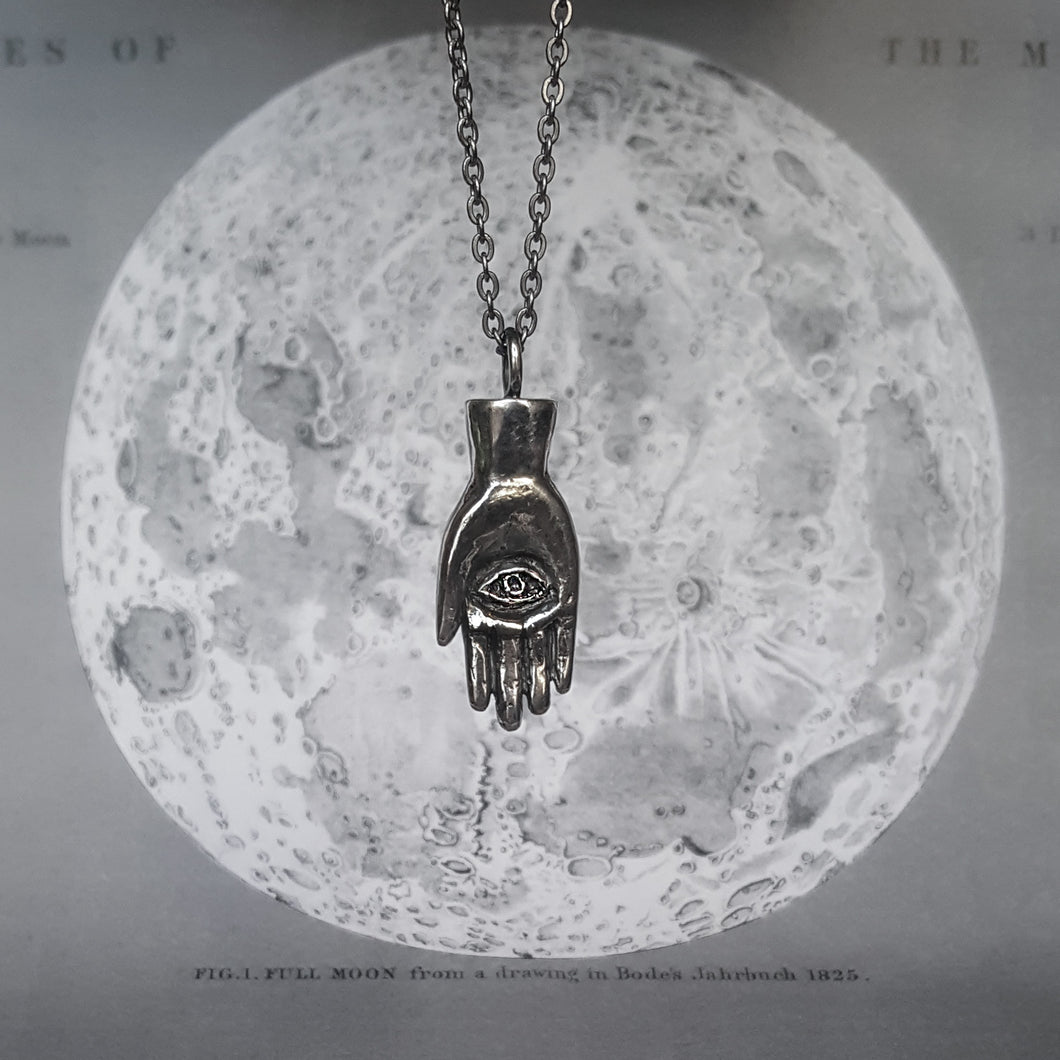 Silver necklace in the shape of a hand with an eye in the palm, also called a hamsa. Shown on a background of the moon. 